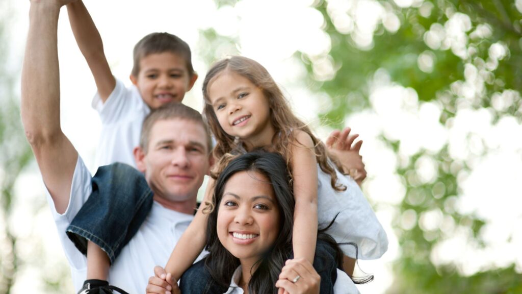 A diverse family wearing white t-shirts with two parents carrying a boy and girl on their shoulders. Blurred green trees are in the background. 