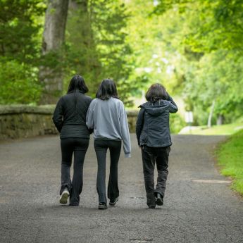 Three individuals with long brown hair and cool weather clothing walking away from the camera on a dirt path with a lush green canopy of trees and foliage in the background. 