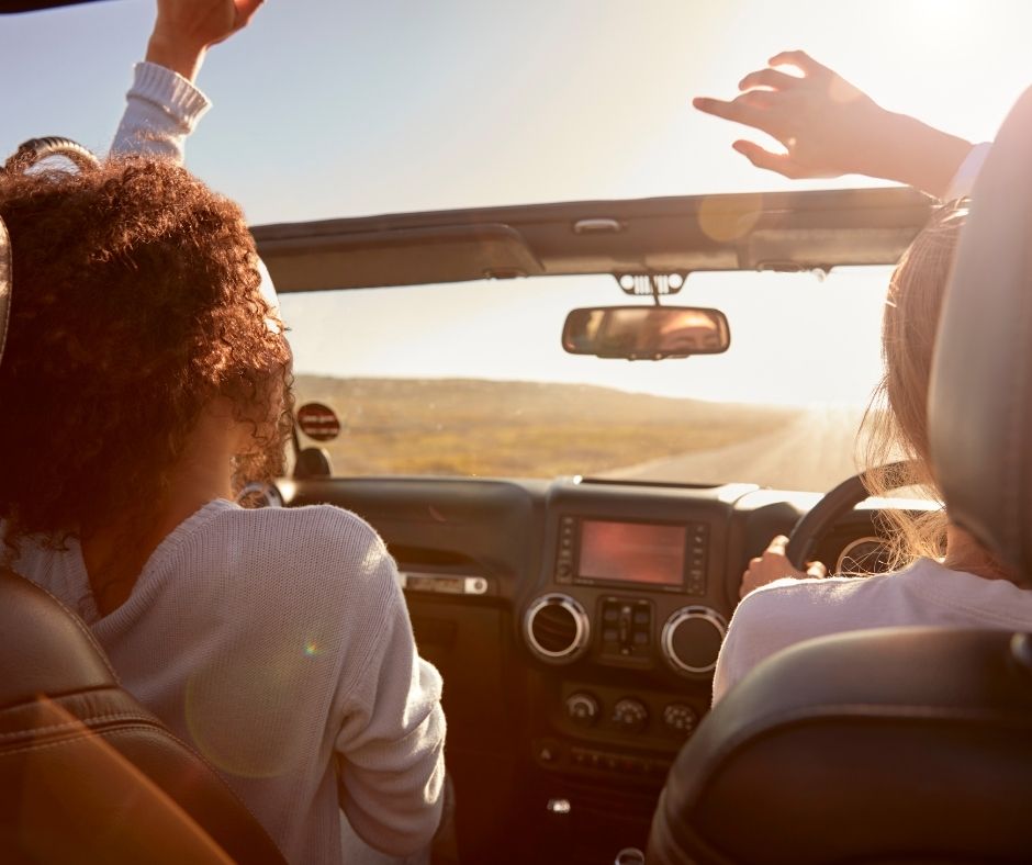 Two women riding in convertible with top down and hands raised. 