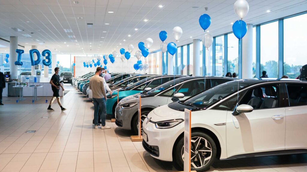 A man is standing in a car dealership looking a row of electric vehicles. There are blue and white balloons throughout the dealership. 