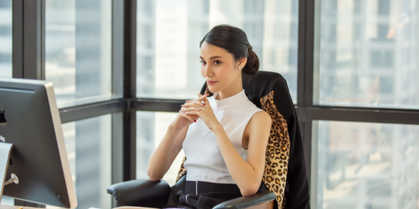 A woman wearing a white mock-turtleneck sits in a chair at her desk in an executive office, with her hands folded and a window behind her.
