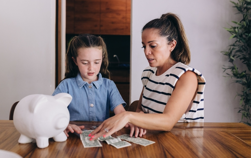 Baby Bonds Colorado A mother teaches her young daughter about saving money at a dining table, with a white piggy bank and cash in front of them.