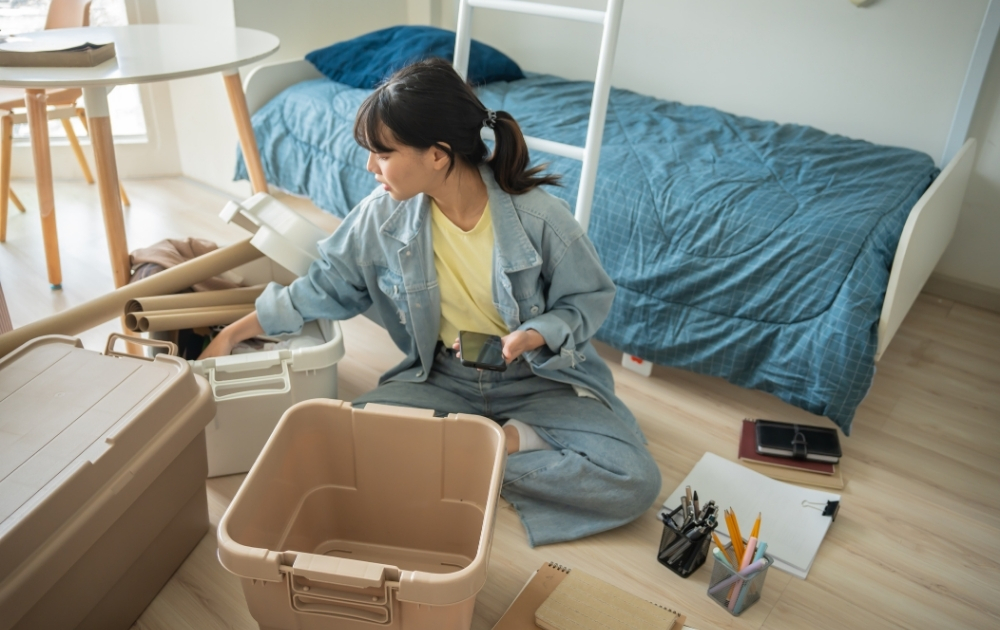 Young woman unpacks storage bins and moving supplies in a bedroom, representing the transition to independent adulthood.