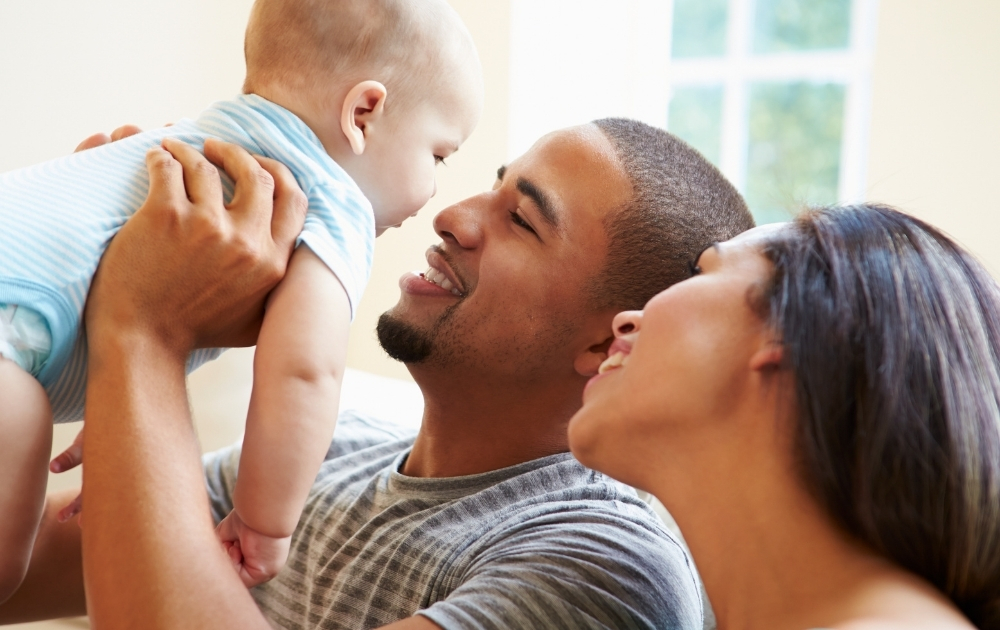 Baby Bonds Colorado Smiling parents spend time with their infant at home as the father lifts the baby in his arms.