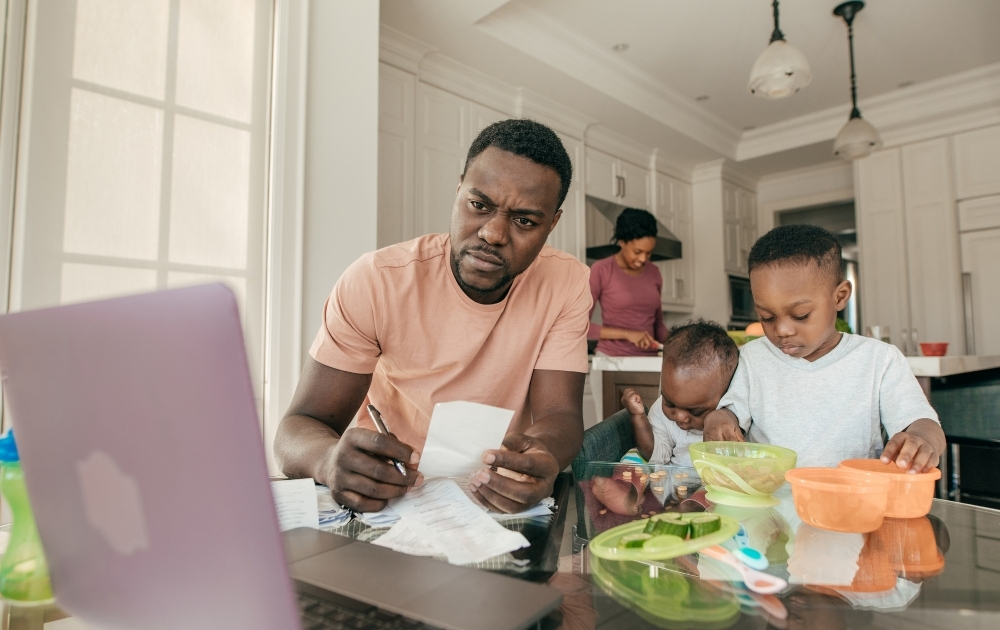 Father reviews household bills at a kitchen table while two young children eat and their mother prepares food in the background.