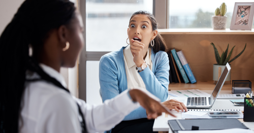 Two women talking in an office, with one woman gesturing in the foreground while the other reacts with a shocked expression, illustrating workplace gossip and the impact of interpersonal communication.