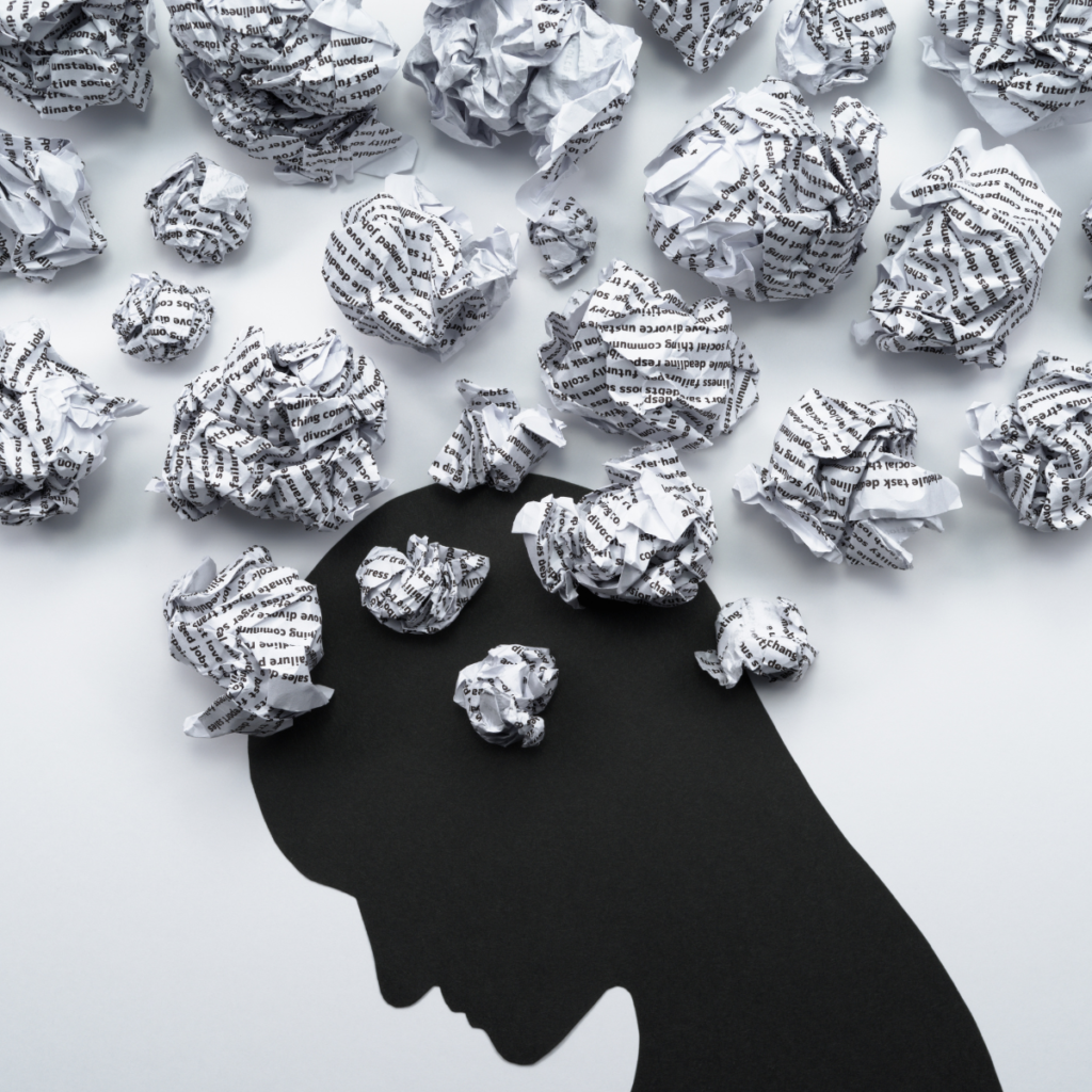 Black silhouette of a person's head against a light background, surrounded by crumpled sheets of paper covered in printed text, symbolizing mental stress, overwhelming information and negative communication.